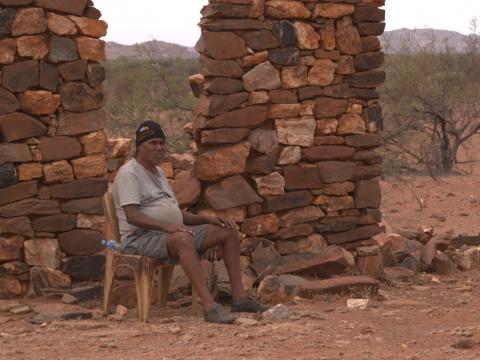 A man sits on a chair in front of a stone wall on a plain in the outback.