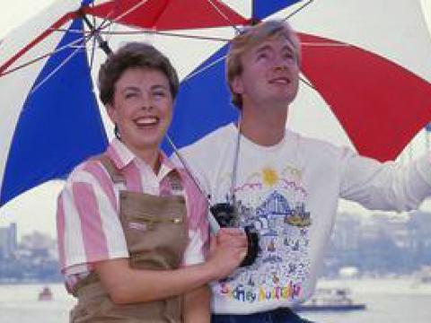 Jayne Torvill and Christopher Dean posing in front of the Sydney Harbour Bridge in the rain with umbrellas.