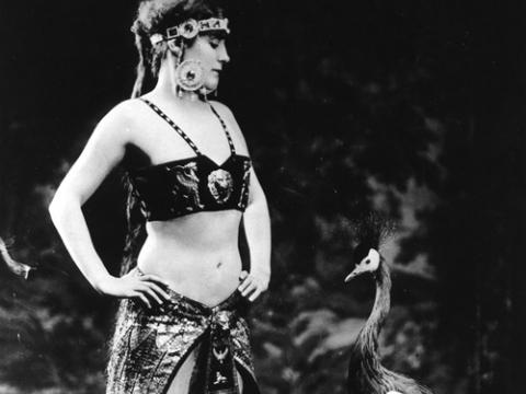 Annette Kellerman stands with her hands on her hips, looking down at a crowned crane bird. Annette is wearing a be-jewelled top, skirt and headdress.