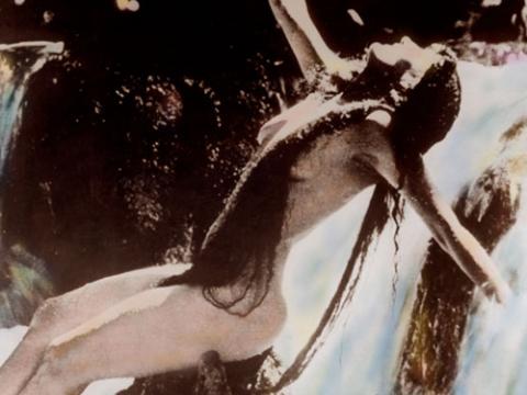 Annette Kellerman in a hand-tinted photograph. She appears to be naked but is covered by her long hair. She is sitting on a rock near a waterfall.