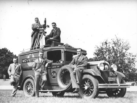 Bill Trerise with a camera on a tripod and one other man on top of Movietone Sound camera car 56, with three other men on the ground.