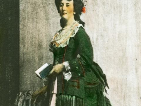 A woman stands in a sitting room holding a book. She is dressed in a late-Victorian era dress with a bustle and has flowers in her hair. The slide is hand-coloured.