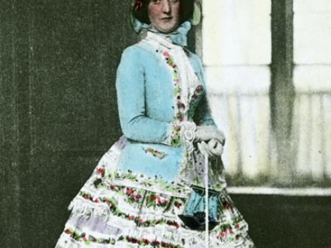 A woman standing near a doorway, wearing a dress from the Victorian era. She has a bonnet, gloves and a parasol. The slide has been hand-coloured.