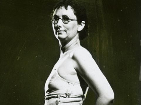 A woman stands looking at the camera. She is wearing spectacles, shoes and a spotted girdle.