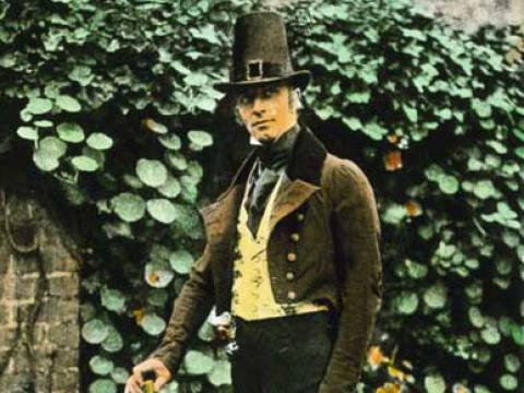 A man stands in front of a garden wall covered in leaves. He's wearing a theatrical costume with hat and waistcoat.