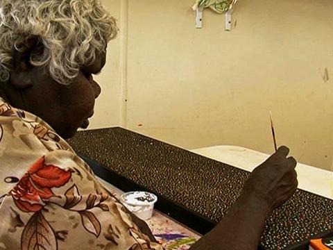 An older Aboriginal woman sitting at a table painting.