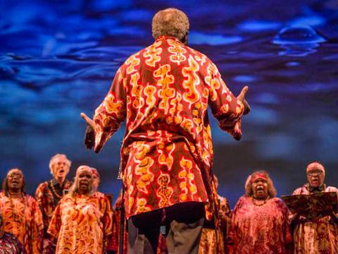 An Aboriginal women's choir being conducted by a man standing in front of them with his back to camera and arms outstretched. 