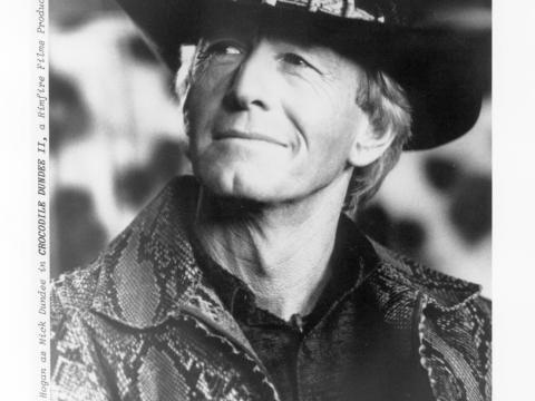 Black and white production still of Mick Dundee (Paul Hogan). He is wearing Mick's trademark hat with crocodile teeth around the band and a crocodile skin jacket.