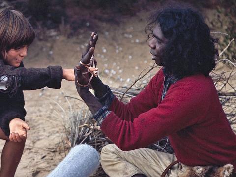 Actors Greg Rowe and David Gulpilil sitting down in the sand, playing with string on the set of Storm Boy. The boom operator with boom and clapper board are visible in the frame