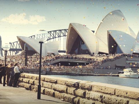 The Sydney Opera House as seen from the Botanical Gardens on its opening day. There are lots of people and colourful balloons. The Sydney Harbour Bridge is in the background