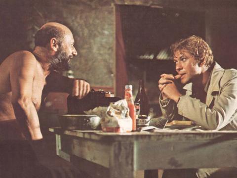 Donald Pleasence sits opposite Gary Bond at a table in his outback hut in a photo from the film Wake in Fright