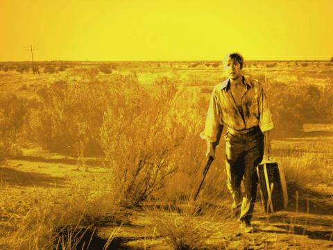 John Grant walks through the harsh yellow light of the outback carrying a suitcase and a gun in an image from the film Wake in Fright
