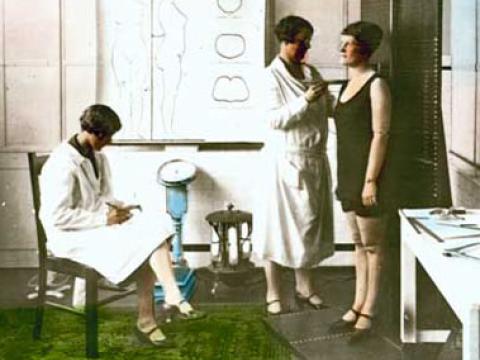 A woman wearing a white coat measures another woman in her underwear. A third woman (also in a white coat) takes notes. They are in a medical-looking room.