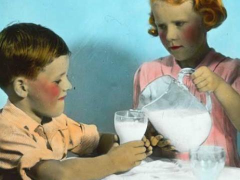 A young girl fills the cup of her smaller companion with milk with a somewhat deadly look in her eye.