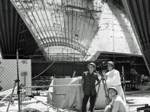 Sails of the Sydney Opera House under construction. A camera crew of three men pose amidst the construction site.