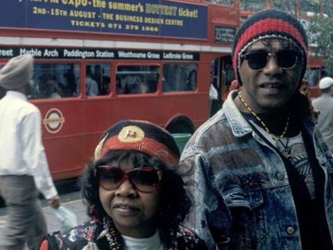 A photograph of Archie Roach and Ruby Hunter standing on a busy street in London. People walk behind them and there is a red double decker bus in the background. 