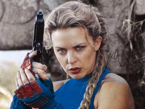 Kylie Minogue poses holding a gun wearing fingerless gloves. Her steely gaze directed to the camera.