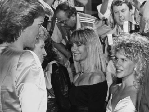Kylie Minogue, Diana Princess of Wales and Olivia Newton-John stand next to one another in this black and white photograph. Kylie is awestruck looking up at Diana who is much taller.