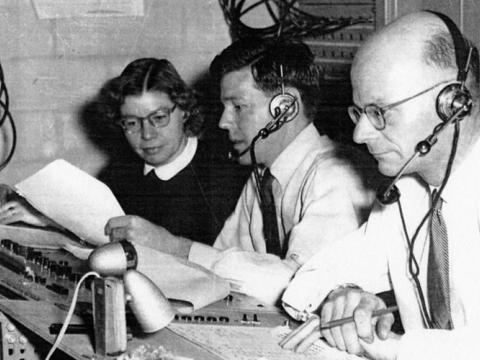 Two men and a woman sit behind a control desk in a black and white photo. There are lots of wires around them. The men wear headsets with headphones.