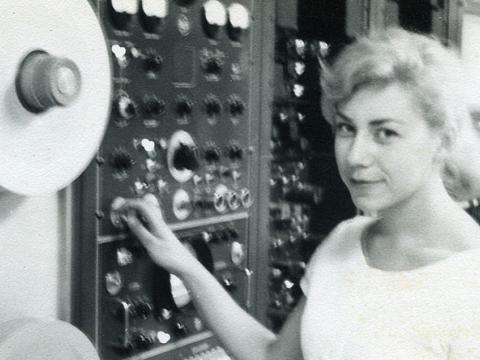 A woman is standing at a deck of videotape machines. She is operating one of the dials as she looks at the camera.