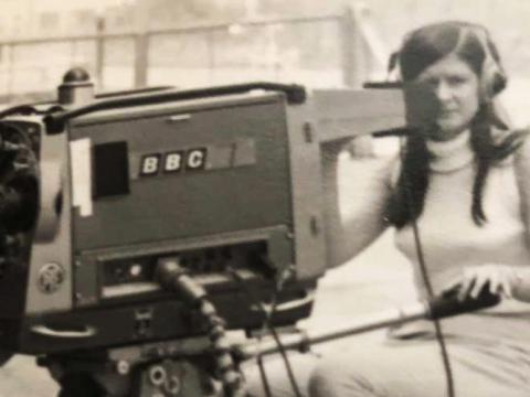 A woman is operating a BBC news camera on a rigging. She is wearing headphones. A man behind her peering into the camera viewfinder.