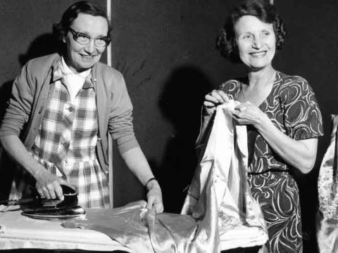 Two women are smiling for the camera. One of them is ironing a piece of clothing on an ironing board and the other is holding some fabric.