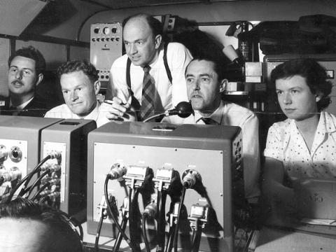 Four men and a woman sit inside an outside broadcast van. There are dials, cords and switches all around them.