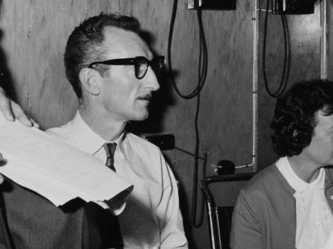 A man and a woman sitting in a studio. They are surrounded by wires and someone holding a script.