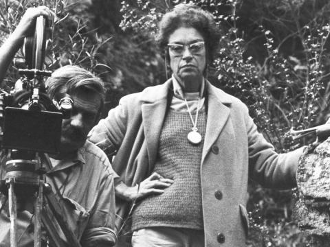 A portrait of two people. A man is holding a film camera on a tripod. A woman is standing with her hand on her hip. They are in a natural setting.