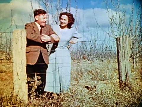 A young couple stand arm in arm by a fence overlooking an orchard, circa 1949