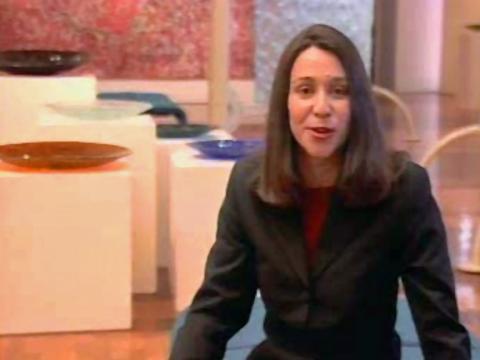 A news reporter sits on a bench in a gallery, behind her are glass artworks.