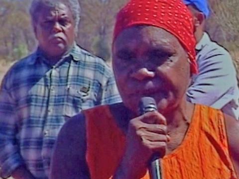 A head and shoulders shot of an Aboriginal elder. She is wearing a bright red head scarf and orange top and is speaking into a microphone.