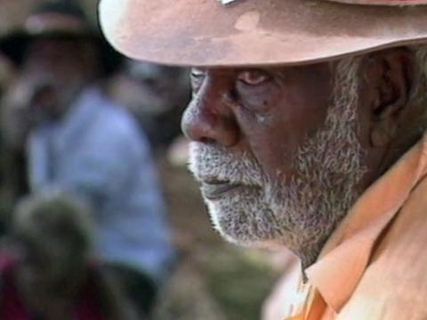 Close up of an elder Aboriginal man with a white beard wearing a hat and a peach-coloured collared shirt. He is pictured in profile with a somber look on his face.