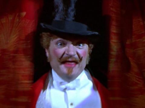 A man in make-up and a top hat looks out from between red velvet curtains