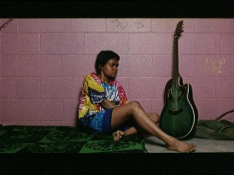 Della (Ursula Yovich) wearing a tie-dyed t-shirt sitting on the floor next to a guitar