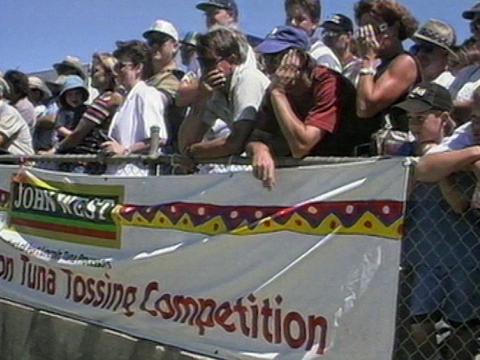 A crowd of people standing behind a fence. In front of the fence is a sign that says "John West Tuna Tossing Competition"