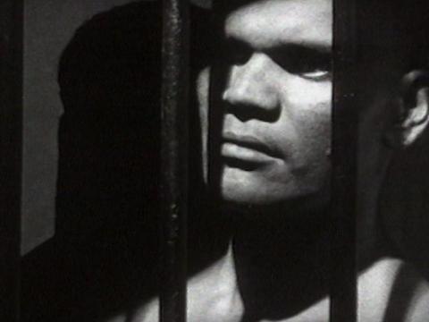 Black and white close up of Paddy (George Djilaynga) looking from behind jail bars