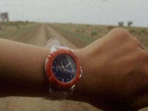 Film still of a red watch on a forearm with a dirt road in the background