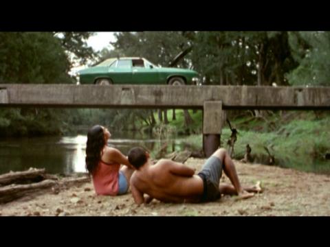 A man and a woman resting on the bank of a river underneath a bridge where a green car is parked