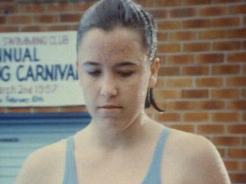 A head and shoulders shot of young girl in swimwear at in front of a brick wall and a swimming carnival sign