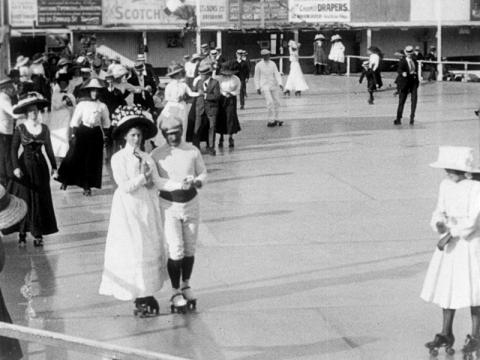 Men, women and children rollerskating on a rink. It is a black and white still and they are wearing clothes from the early 1900s.
