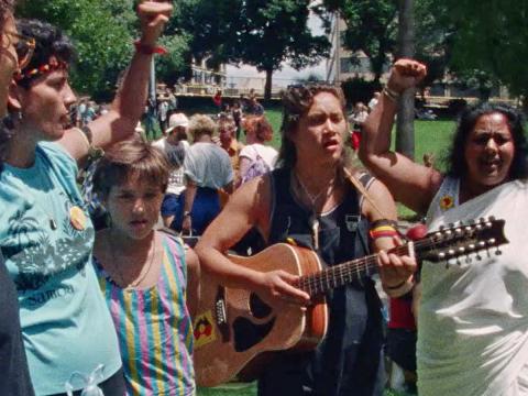Four Indigenous women and one child standing side by side in a crowded park. One is playing guitar and the others are singing with one arm raised in a fist as a symbol of strength.
