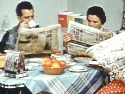 A man and woman sit down at a breakfast table reading newspapers.