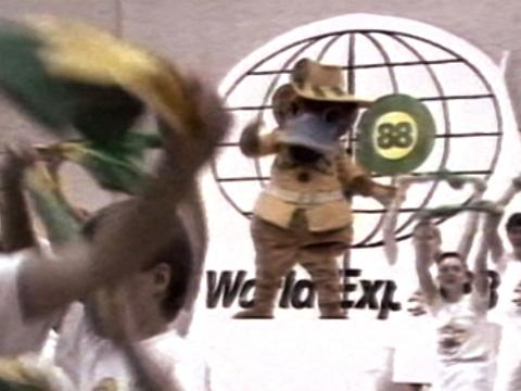 A person dressed as a giant platypus wearing a safari jacket and akubra hat is dancing on a stage. The words World Expo 88 are in the background. Children dance around nearby.