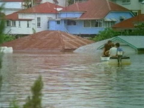 Flood waters cover entire houses, with only the roofs showing above the tide. Two people are in a boat going down what is probably a road underneath the flood water.