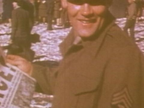 A man in army uniform is smiling and holding a newspaper with the headline 'Peace'. He is standing in a crowd of people.