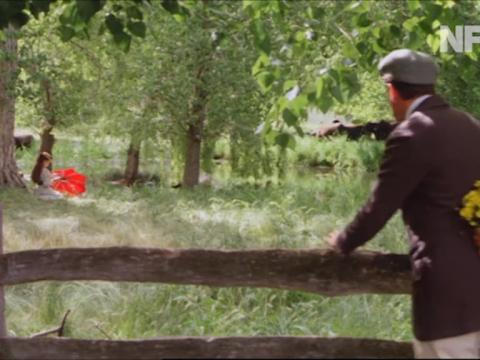 Would-be suitor Frank Hawden (Robert Grubb) stands at a fence with flowers behind his back watching Sybylla (Judy Davis) who sits under a tree by the lake