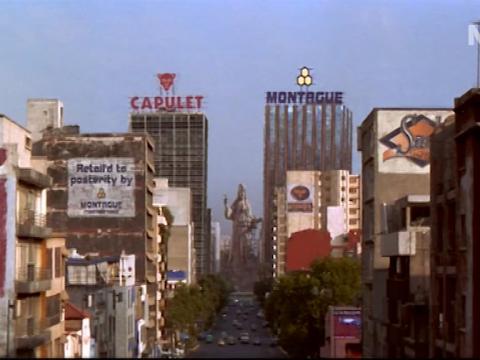 Shot of a city, two buildings dominate the image, one with a sign saying 'Capulet' and the other 'Montague'