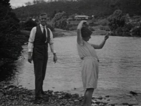  A man stands looking at the camera as a woman combs her hair looking in a mirror by the riverbank