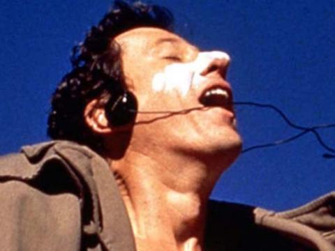Head and shoulders shot of Geoffrey Rush with white sun cream across his nose and wearing headphones, jumping on a trampoline against a bright blue sky, with his eyes closed and mouth open in ecstasy